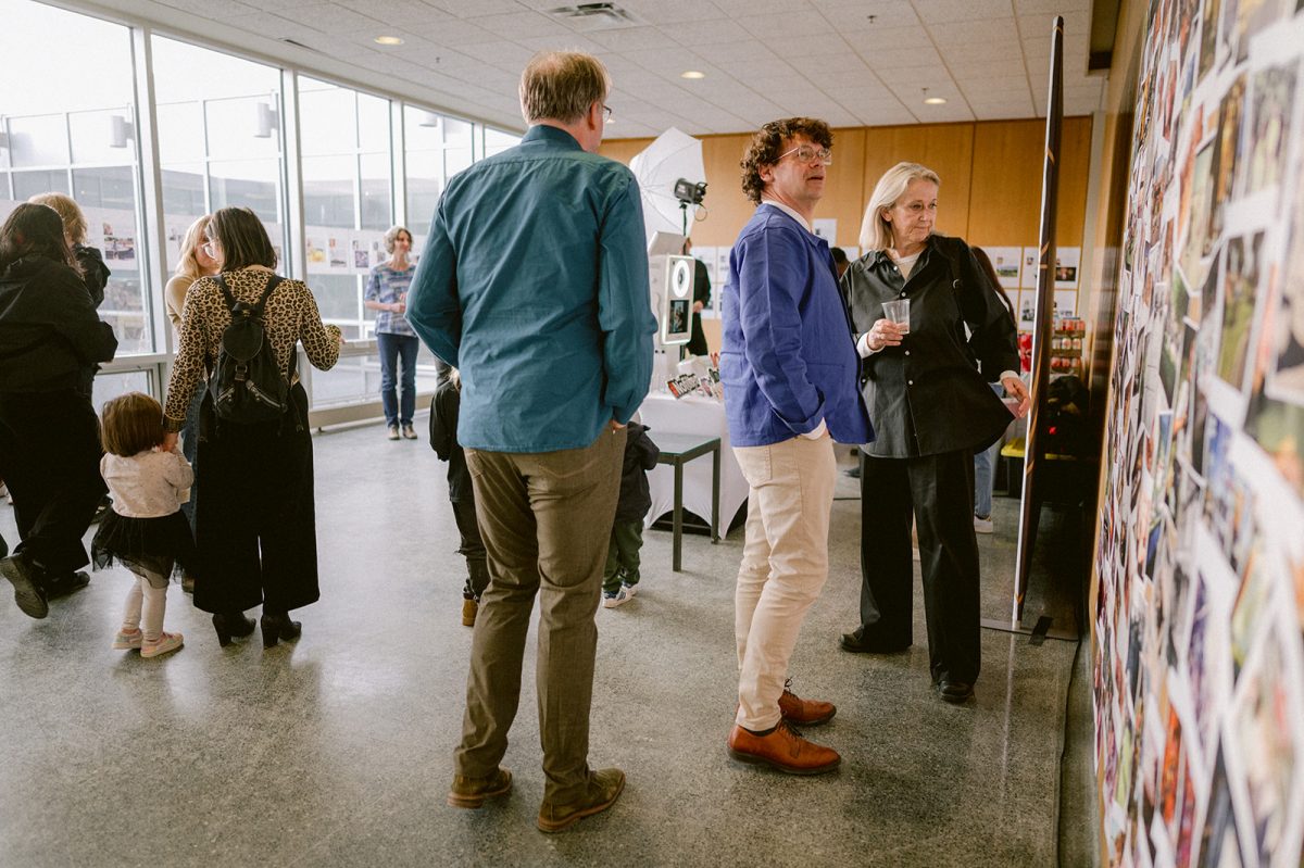 A man and woman look at photos displayed on the wall, while other people mill about in the background.