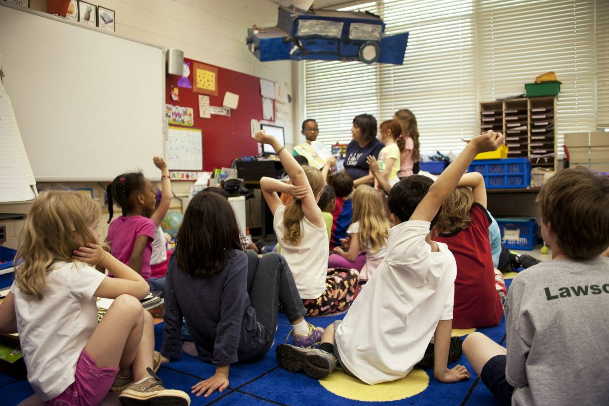 school children were seated on the floor before a teacher at the front of the room, who was reading an illustrated storybook, during one of the scheduled classroom sessions.