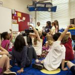 school children were seated on the floor before a teacher at the front of the room, who was reading an illustrated storybook, during one of the scheduled classroom sessions.