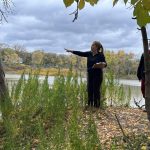 Three people standing on a riverbank with trees and water around them.