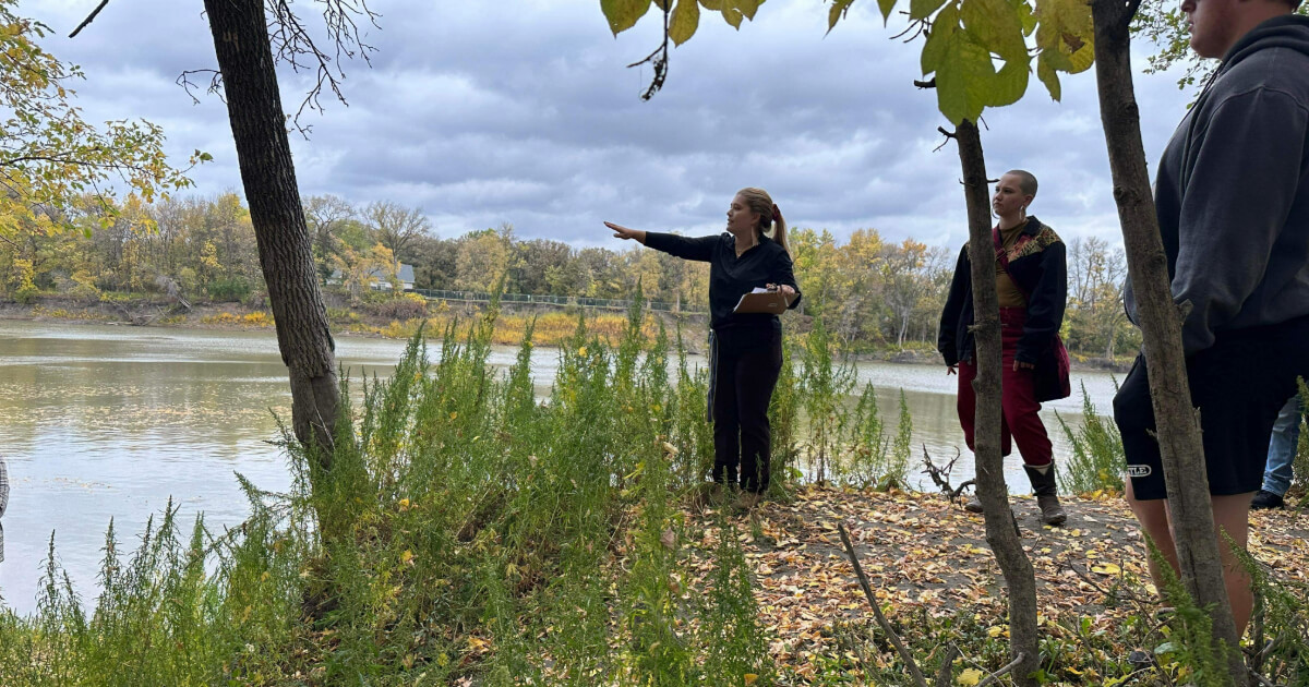 Three people standing on a riverbank with trees and water around them.