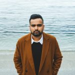 Nabil Iqbal stands in front of a body of water, his hands in his pockets, looking at the camera.
