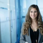Woman with long brown wavy hair, wearing a grey suit jacket and v-neck black shirt.
