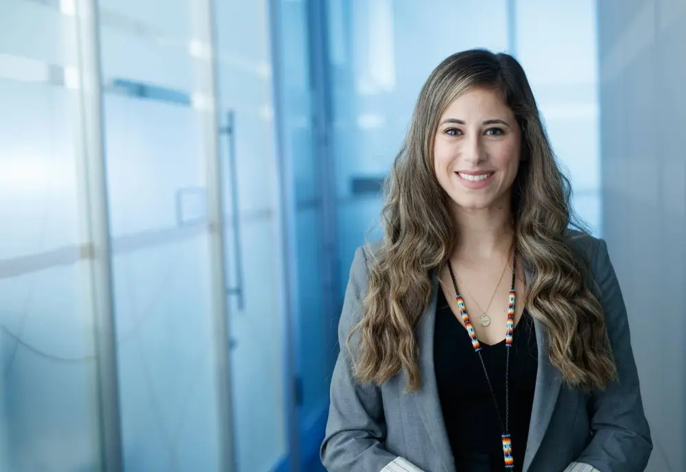 Woman with long brown wavy hair, wearing a grey suit jacket and v-neck black shirt.