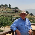 haskel greenfield, a man wearing a blue button up shirt and a beige hat, pictured on a wood deck with scenic hills behind in the distance.