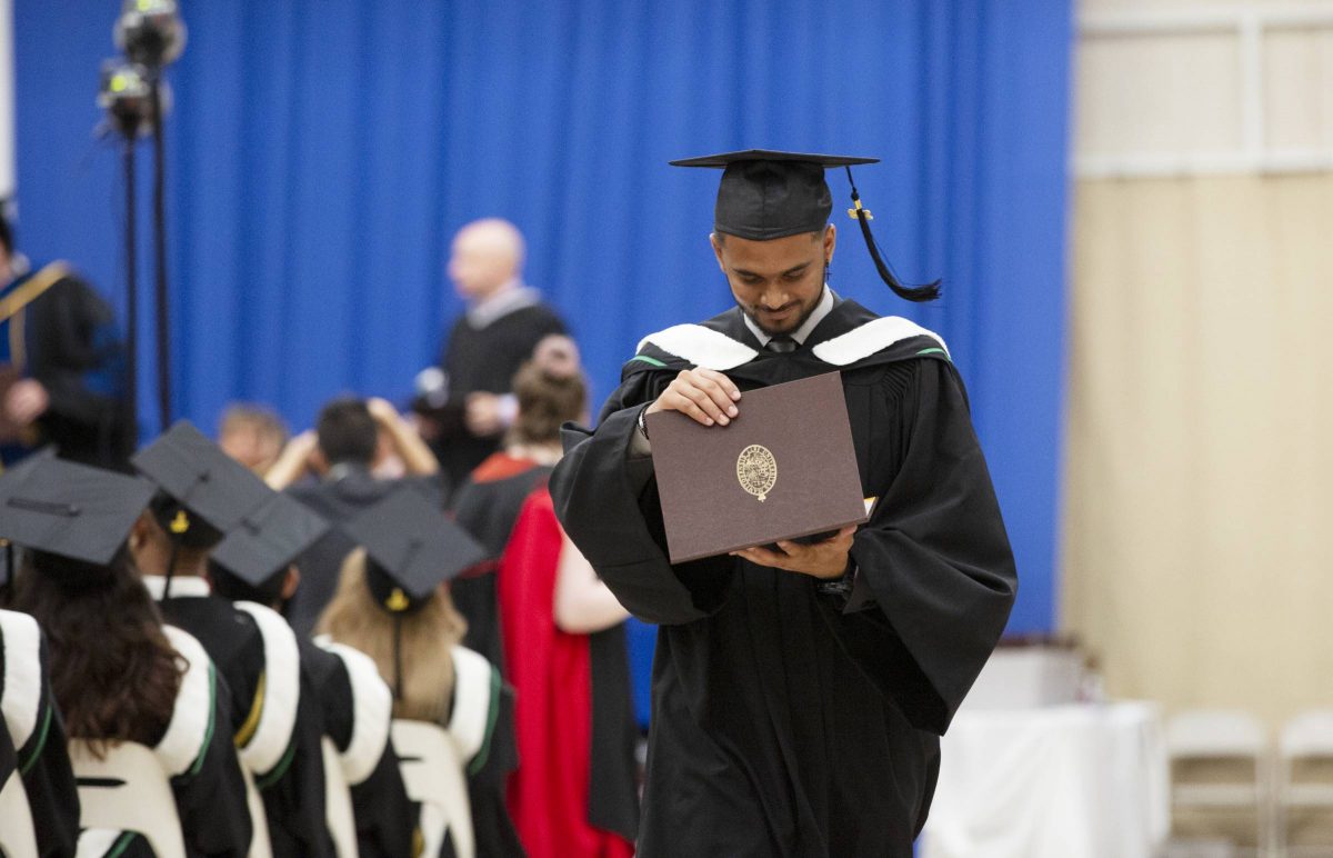 A UM graduate dressed in a cap and gown walks away from the stage at Convocation. They are holding their open parchment in their hands and smiling down at it, celebrating their achievement.