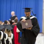 A UM graduate dressed in a cap and gown walks away from the stage at Convocation. They are holding their open parchment in their hands and smiling down at it, celebrating their achievement.