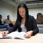 An Asian female student studies in a student lounge, with other students meeting behind her.