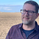 Man with short salt and pepper hair and beard, wearing black rectangular frame glasses. He is wearing a purple button down shirt with a dark blow shirt underneath. He is standing in front of a farmers field.