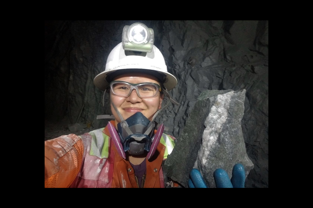 Woman in a white safety helmet with safety goggles, a breathing mask and orange protective clothing. She is standing in a mine holding a large chunk of stone.