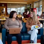 Two students with laptops sit on a couch in a student lounge. They are smiling at each other.