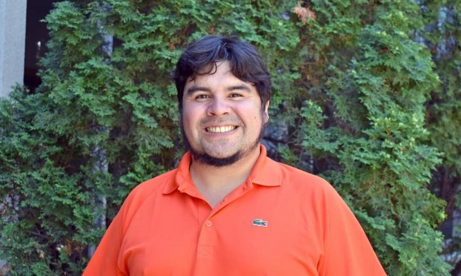 Kyle Bobiwash smiles wide in front of cedar bushes.