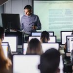 Professor at laptop in front of of projection screen, lecturing to students