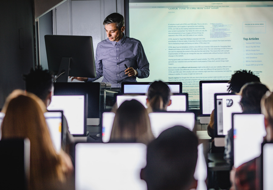 Professor at laptop in front of of projection screen, lecturing to students