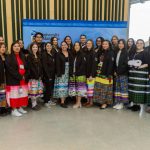 A group of students, many in ribbon skirts, stand together in a semi circle.