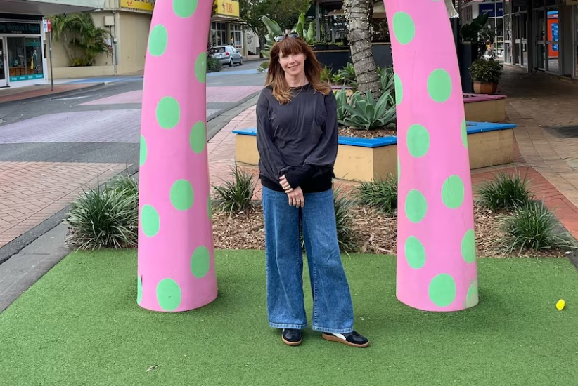 woman with should length brown hair, brown bangs. Wearing a long sleeve black sweatshirt, blue jeans and black sneakers. standing outside under a pink with green polka dots arch.