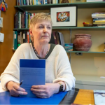 Woman with short blonde hair wearing a cream colour v-neck sweater holding a blue book and sitting in front of a book case.