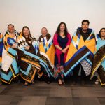 A group of students, staff and faculty members stand in front of a white wall, wrapped in Star Blankets.