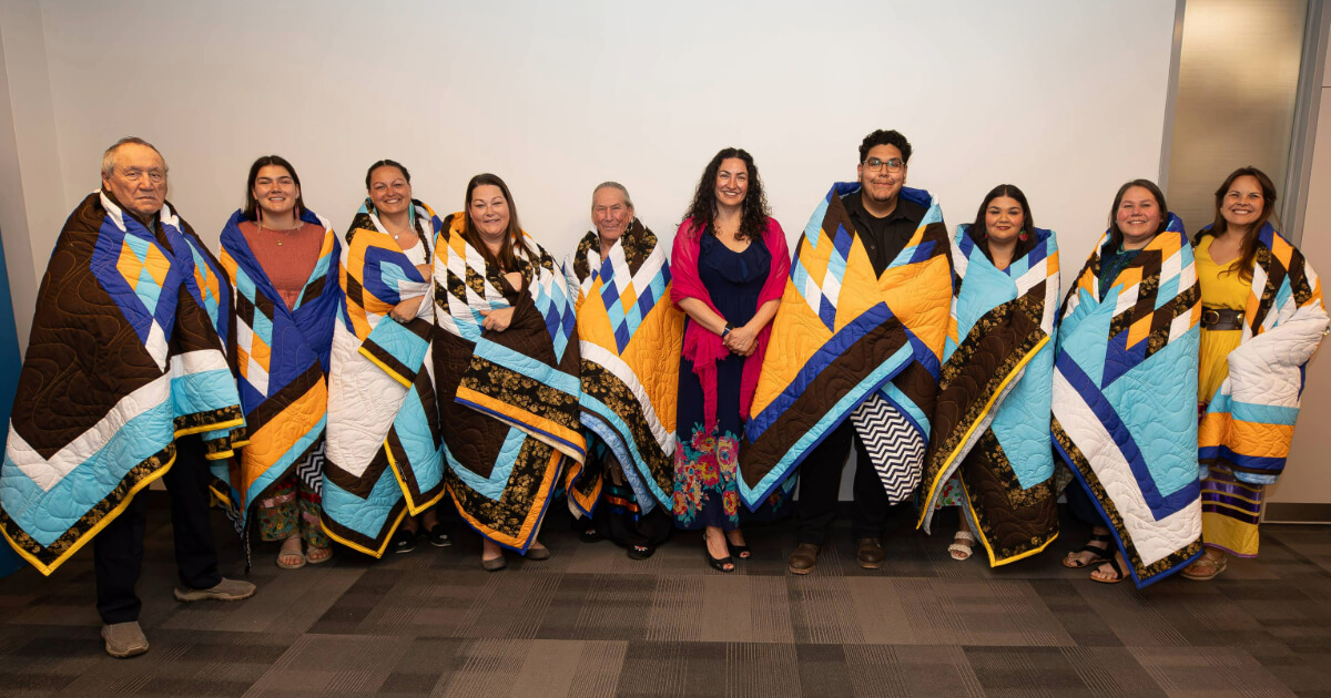 A group of students, staff and faculty members stand in front of a white wall, wrapped in Star Blankets.
