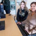 Two female students sit together at a computer, smiling at the camera.