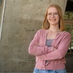 Female wearing glasses standing with arms crossed against a backdrop of a cement wall.