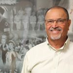 Peri Venkatesh stands in front of a black and white collage that shows the history of nursing in Manitoba.