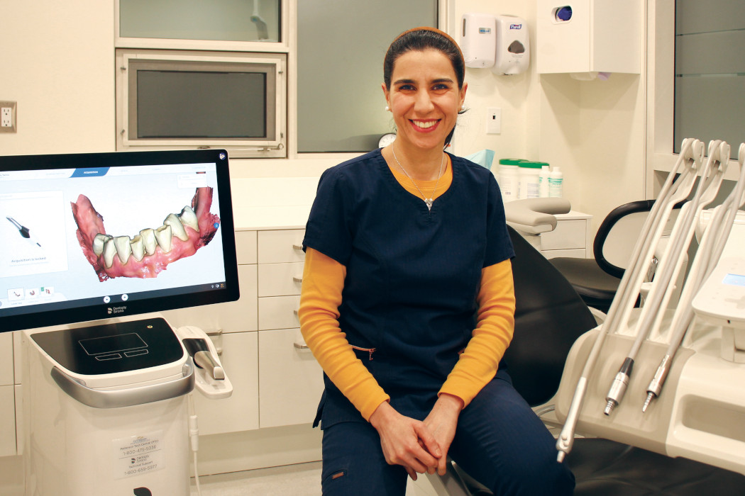 Dr. Hoda Hosseini sits on a dental chair. A 3D image of teeth and gums is on a screen next to her.