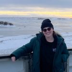 Dr. Emma Anderson poses for a photo wearing winter clothing. A landscape of snow and ice is in the background, with the sun at the horizon.