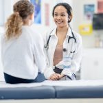 A female doctor talks with a little girl about her health during a medial appointment. She is dressed semi-casually and has a stethoscope around her neck.