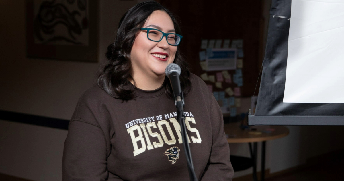 A student with long dark hair and glasses wearing a UM Bisons crewneck sweater stands at a microphone, smiling.