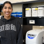 Shubhneet Thind, smiling at the camera, standing in a biochemistry lab.