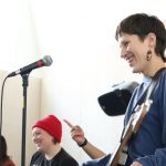 Person with short hair and blue sweater stands in front of a mic on a mic stand. They are holding a guitar. There are two other people to their left, also smiling and with mic's on mic stands.