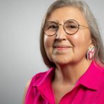 Headshot photo of Pahan PteSanWin. She is wearing glasses, facing the camera and smiling. She has long silver hair and is wearing a pink shirt, set against a grey background.