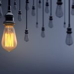 A bunch of lightbulbs hanging from the ceiling, all of them off except one lone lightbulb is lit, standing out from the gray background