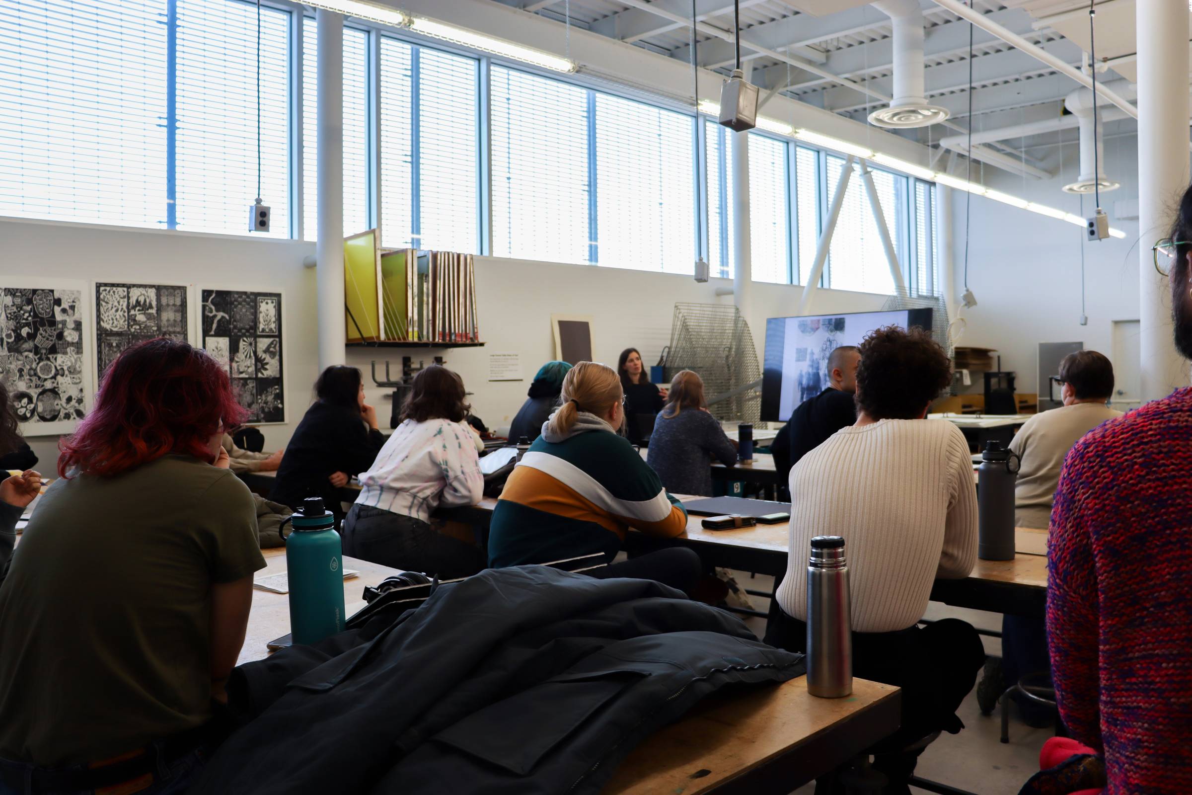 A group of students seated at wooden tables in a well-lit art studio with high ceilings and large windows. They are attentively watching a presenter at the front, who is gesturing towards a large screen displaying visuals. Various art supplies, tools, and racks are visible in the background, along with artwork displayed on the walls.