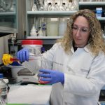 Dr. Inna Rabinovich-Nikitin is in her lab wearing a white lab coat. She is pouring a red liquid from one container to another.