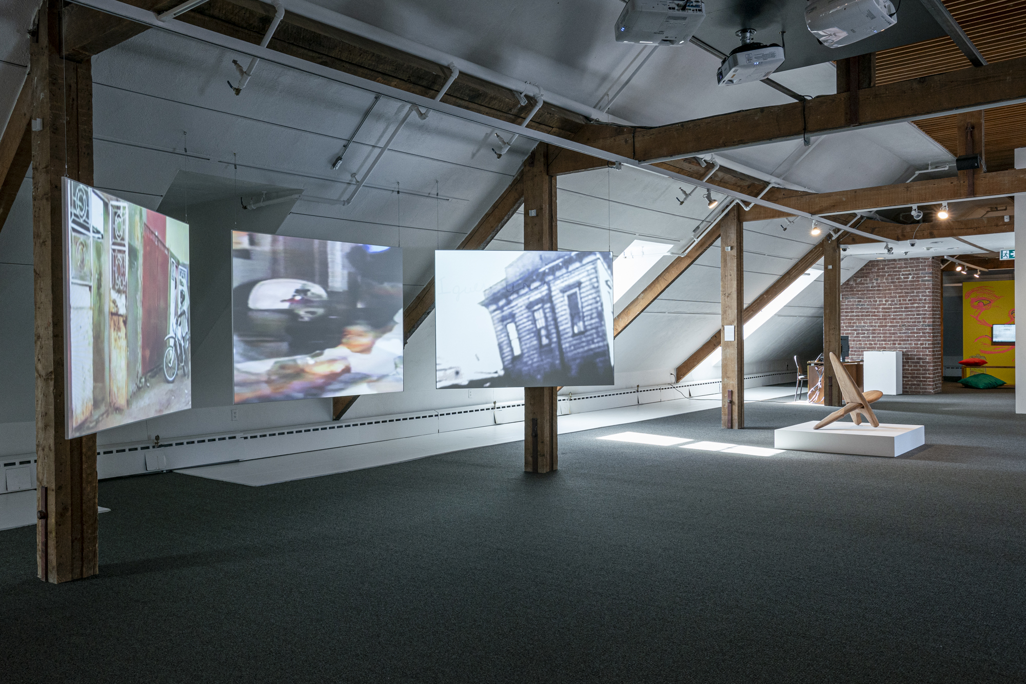 An art installation in a gallery featuring three suspended projection screens displaying video imagery, wooden beams, and a sculptural wooden chair on a white platform.