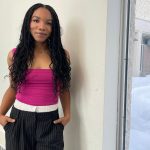 Female student with long black hair, wearing a pink tank top standing in front of a window with both hands in her pockets.