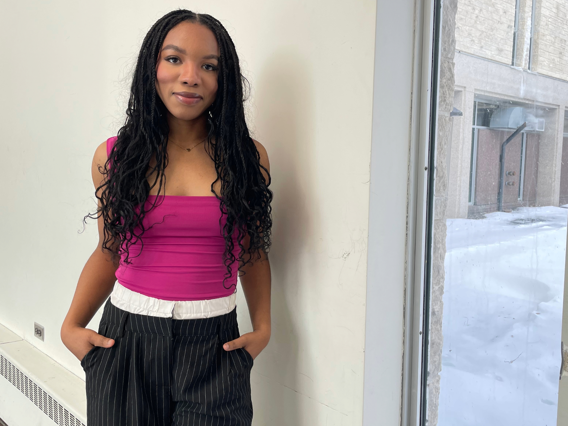 Female student with long black hair, wearing a pink tank top standing in front of a window with both hands in her pockets.