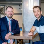 Two men sitting at a restaurant table, looking into the camera and smiling.