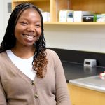 A student smiling at the camera, sitting in a chemistry lab.