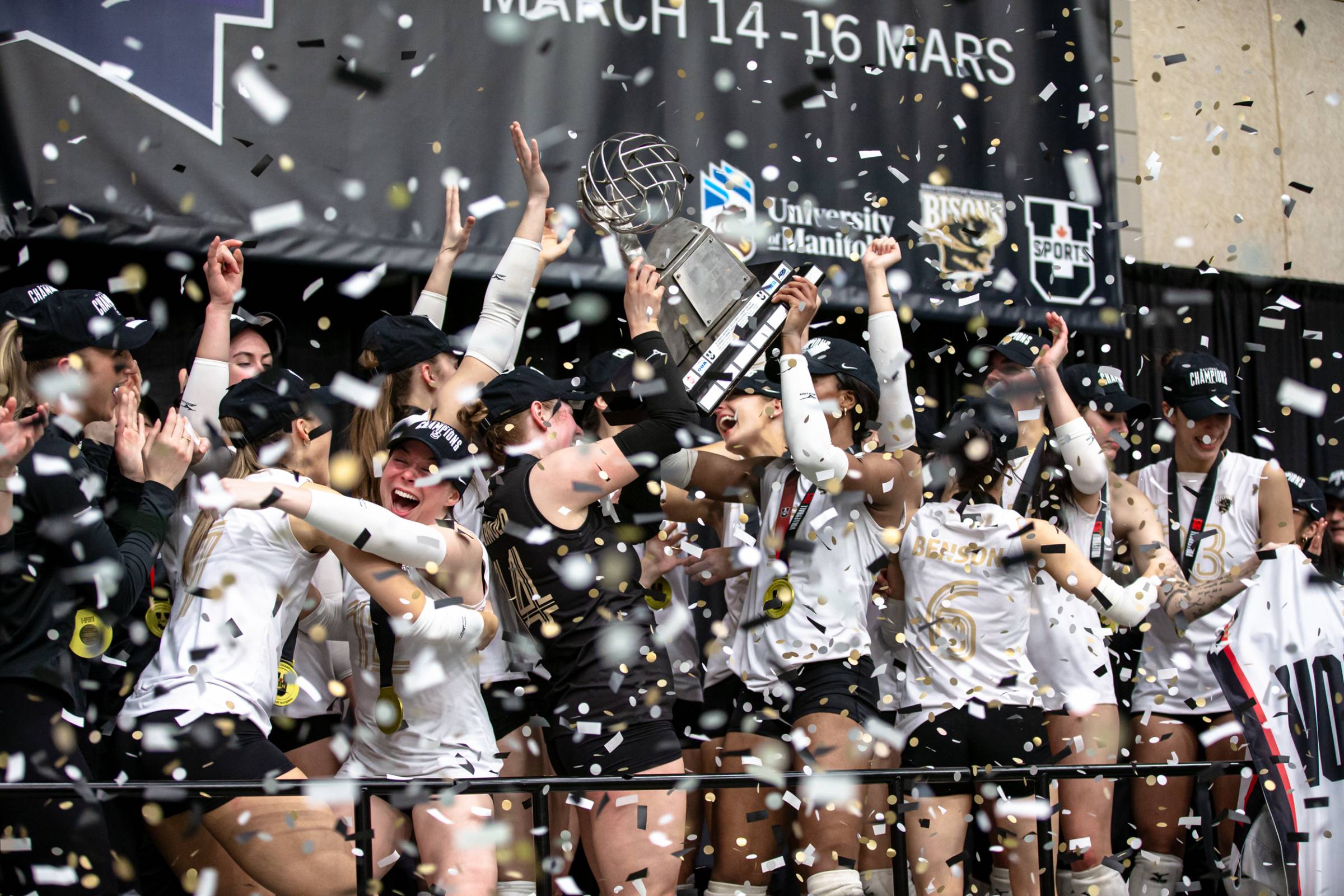 "University of Manitoba Bisons women’s volleyball team celebrates their 2025 U SPORTS National Championship victory. Players, wearing championship hats and medals, cheer and raise the championship trophy high as gold, black, and white confetti rains down. A University of Manitoba and Bisons-branded banner hangs in the background, highlighting the team’s triumphant moment.