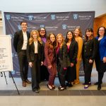 A group of nine people stand in front of a banner and poster for the Canadian National Negotiation Competition March 7 - 8, 2025 at Windsor Law school.