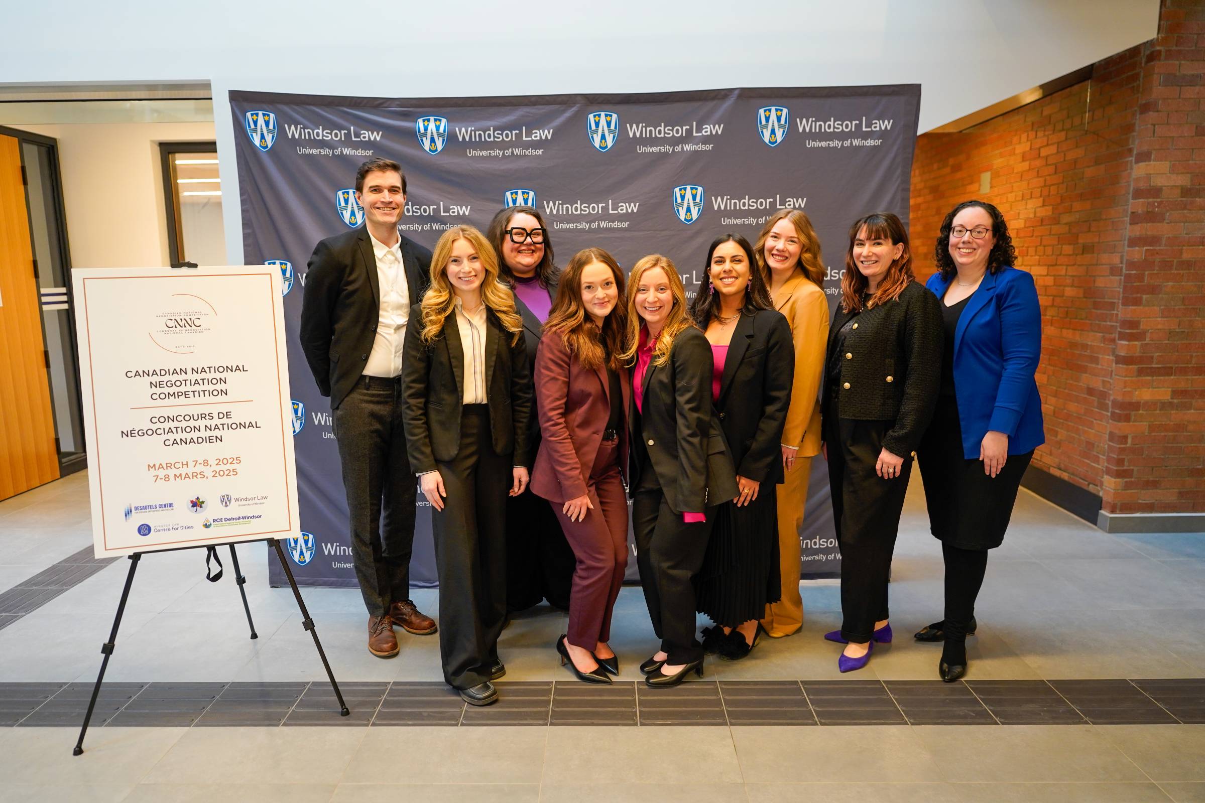 A group of nine people stand in front of a banner and poster for the Canadian National Negotiation Competition March 7 - 8, 2025 at Windsor Law school.