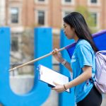 2024 Preparing for University orietation volunteer with a flag infront of the blue UM letters and the admin buiding in the foreground