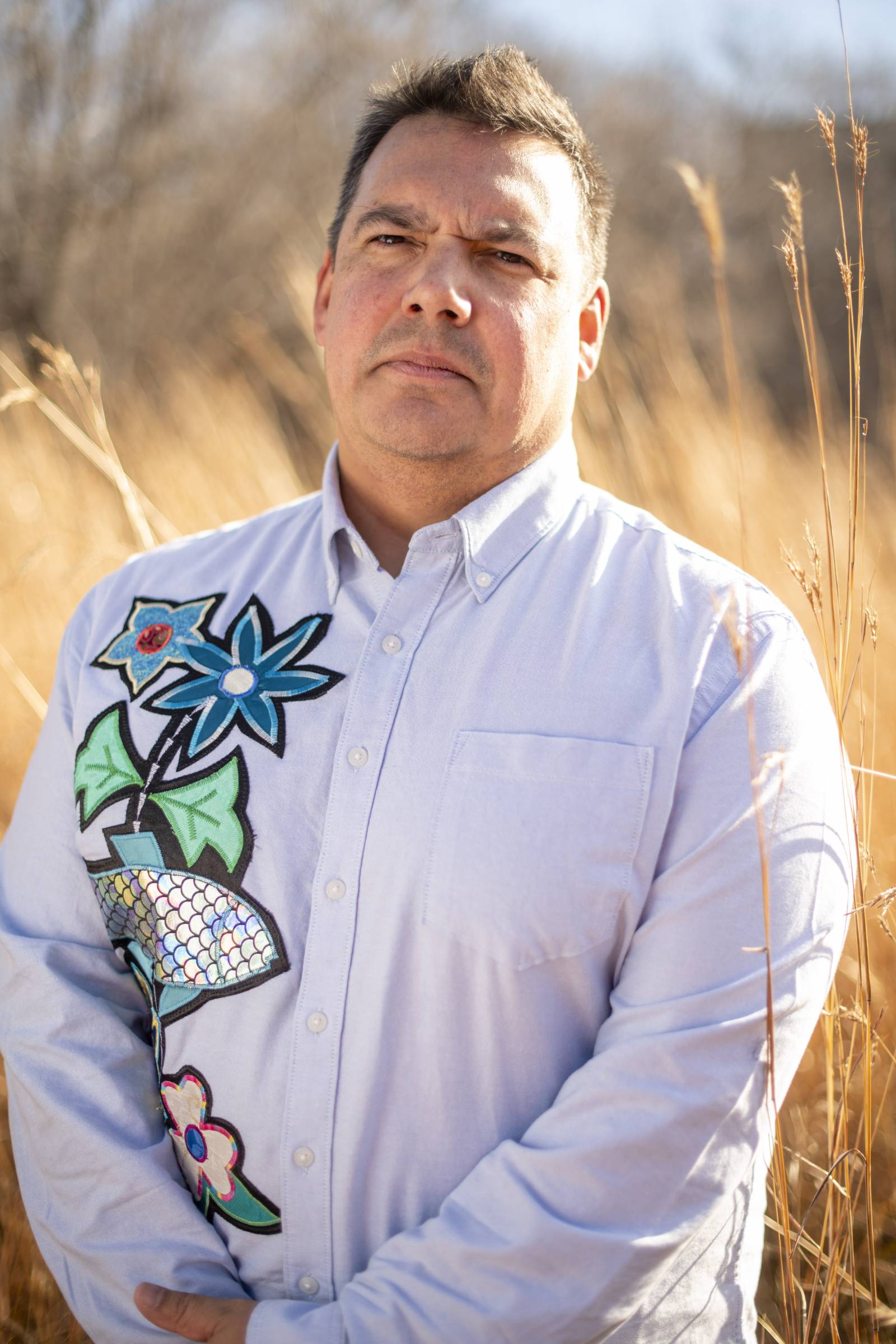 Professor Niigaan Sinclair standing outside in front of a field of tall grasses.