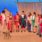 A group of ten actors on stage, dressed in colonial costumes, standing in a semi circle.