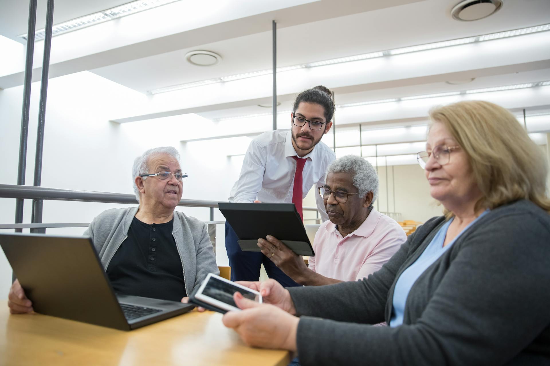 An instructor is standing as he meets with three older adult learners seated in a classroom while they look at their tablets during the class.