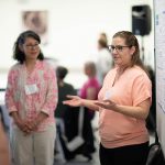 Instructor presents her research, standing in front of a poster display.