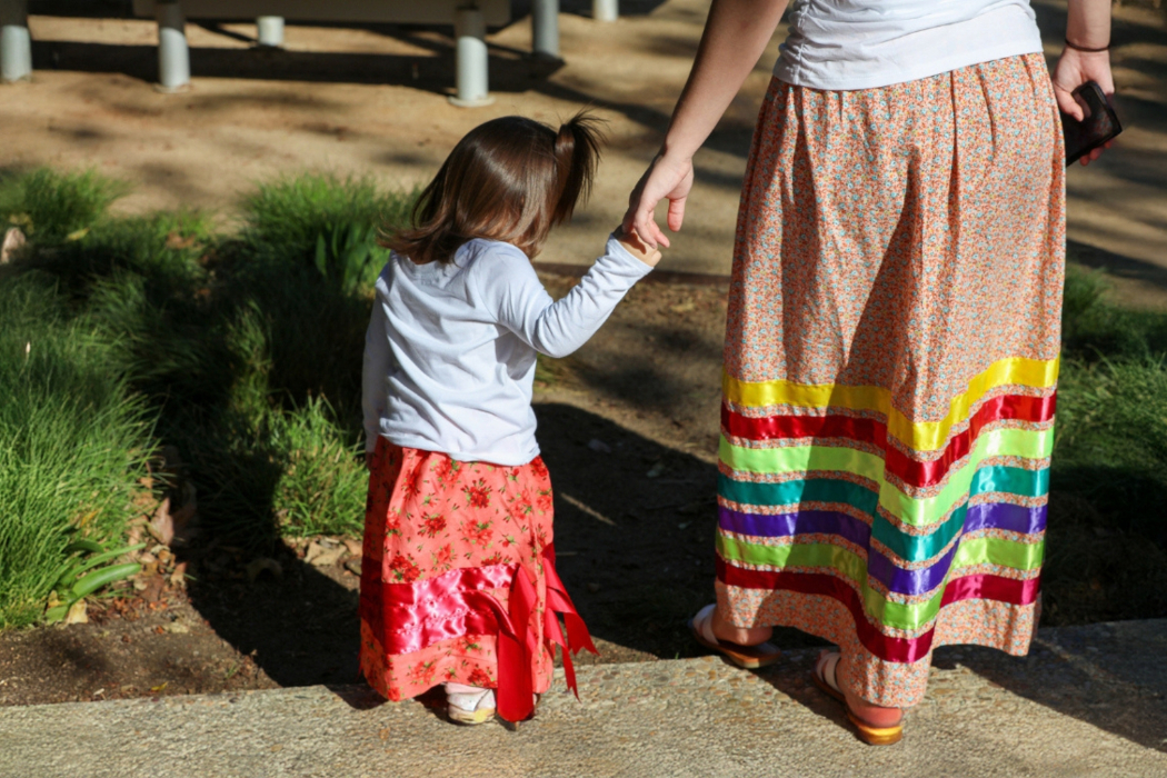 An adult is holding a child's hand. They are wearing ribbon skirts.